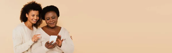 African american adult daughter pointing with finger to smartphone in hands of middle aged mother on beige background, banner — Stock Photo