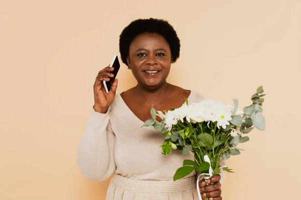 Sonriente mujer afroamericana de mediana edad hablando por celular y sosteniendo ramos de flores sobre fondo beige — Stock Photo