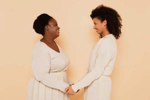Happy african american adult daughter and middle aged mother holding hands and looking at each other on beige background — Stock Photo