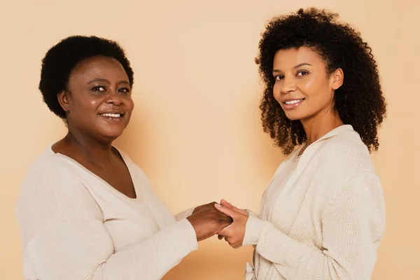 Sorridente afroamericana figlia adulta e madre di mezza età che si tiene per mano su sfondo beige — Foto stock