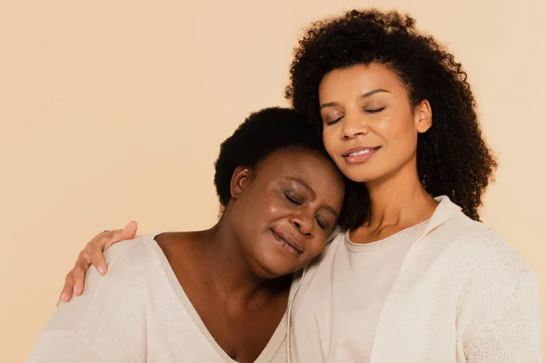 Calma afroamericana hija adulta y madre de mediana edad abrazo con los ojos cerrados aislados en beige — Stock Photo