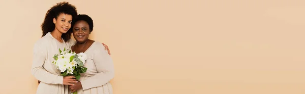 African american adult daughter and middle aged mother standing with bouquet of flowers on beige background, banner — Stock Photo