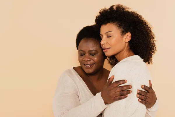 Retrato de hija adulta afroamericana y madre de mediana edad abrazándose con los ojos cerrados aislados en beige - foto de stock