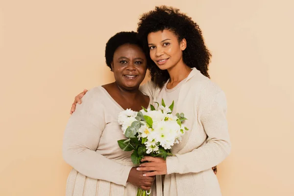African american adult daughter and middle aged mother standing with bouquet of flowers isolated on beige — Stock Photo