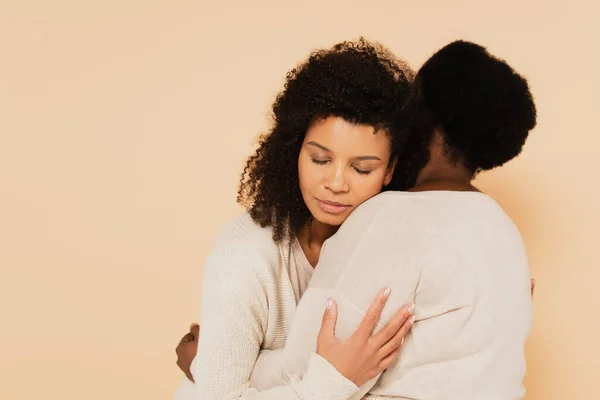 African american adult daughter with closed eyes embracing with middle aged mother isolated on beige — Stock Photo