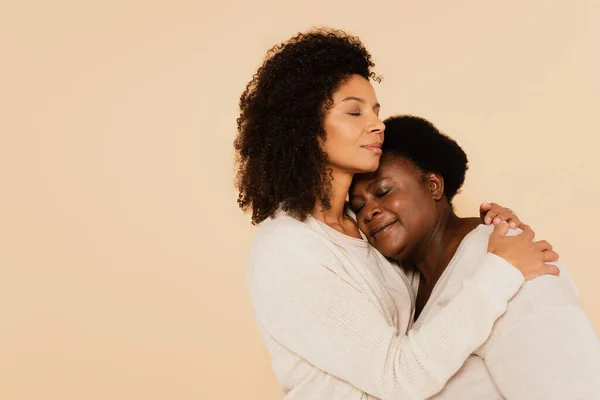Afro-americano adulto filha abraçando meia-idade mãe com olhos fechados isolado no bege — Fotografia de Stock