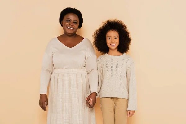 African american grandmother and granddaughter holding hands in pastel clothes on beige background — Stock Photo