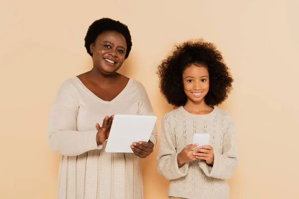 Afrikanisch-amerikanische Enkelin mit Handy und Großmutter mit Tablet schauen in die Kamera auf beigem Hintergrund — Stockfoto