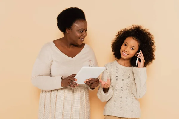 Afro-americana neta falando no celular perto da avó com tablet em mãos no fundo bege — Fotografia de Stock