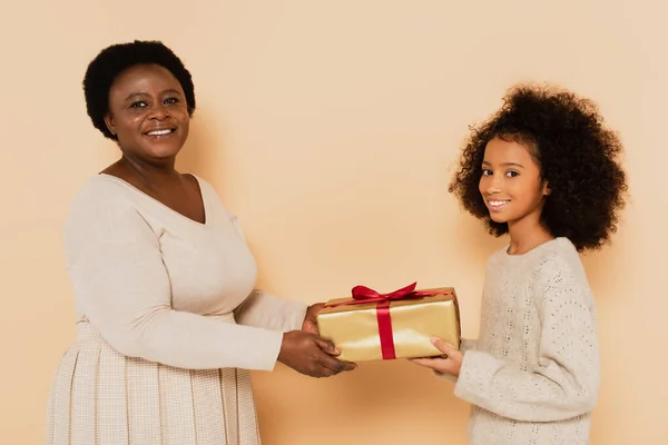 Emocionado afroamericano nieta y abuela sosteniendo caja de regalo y mirando a la cámara en fondo beige - foto de stock