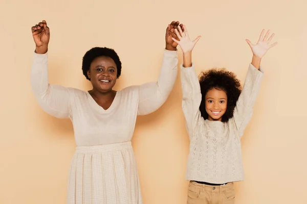 Excited african american granddaughter and grandmother holding hands up on and showing palms on beige background — Stock Photo