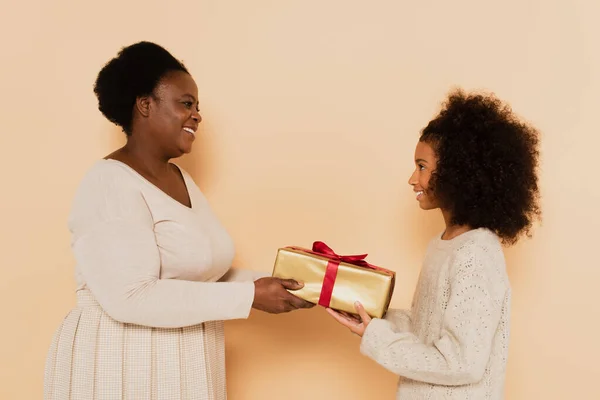 Vue de côté de sourire afro-américaine petite-fille et grand-mère tenant boîte cadeau sur fond beige — Photo de stock