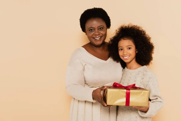 Portrait of smiling african american granddaughter and grandmother holding gift box isolated on beige — Stock Photo