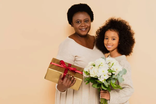 Smiling african american granddaughter and grandmother holding gift box and bouquet of flowers on beige background — Stock Photo