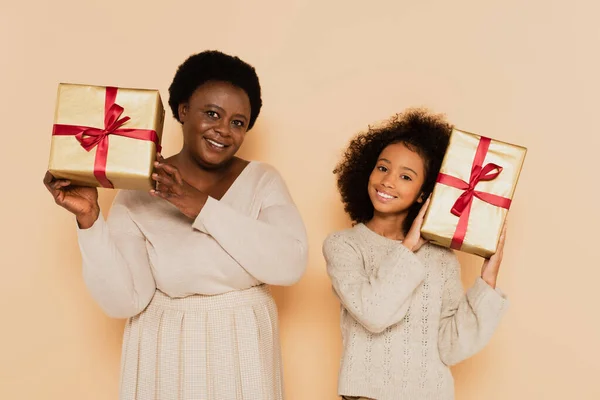 Happy african american granddaughter and grandmother holding gift boxes near heads on beige background — Stock Photo