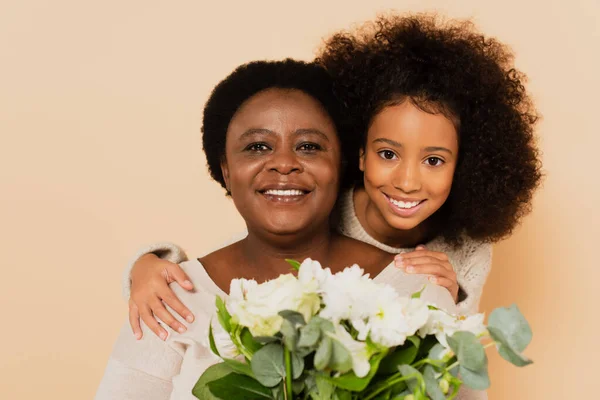 Happy african american grandmother and granddaughter with bouquet of flowers isolated on beige — Stock Photo