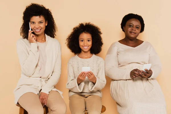 Afro-américaine mère parlant sur téléphone portable près de fille et grand-mère avec smartphones dans les mains sur fond beige — Photo de stock