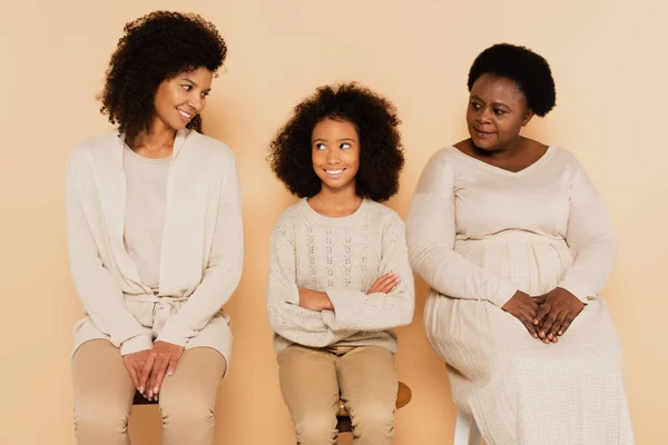 Nieta afroamericana sentada con los brazos cruzados entre la abuela y la madre sobre fondo beige - foto de stock