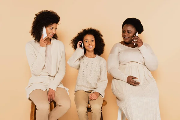 African american grandmother, daughter and granddaughter sitting and speaking on cellphones on beige background — Stock Photo