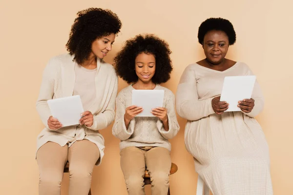 Hija afroamericana, nieta y abuela sentadas y mirando tabletas sobre fondo beige — Stock Photo
