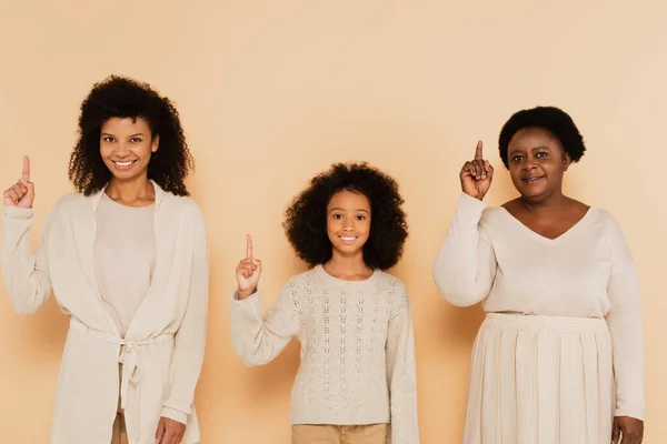 African american daughter, granddaughter and grandmother pointing up with fingers with idea gestures on beige background — Stock Photo