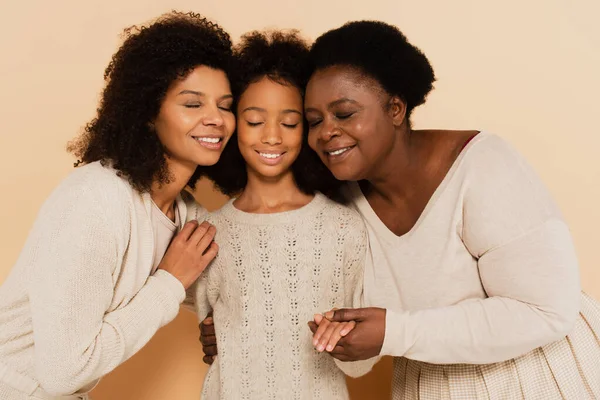 African american daughter, granddaughter and grandmother hugging cheeks to cheeks and holding hands on beige background — Stock Photo
