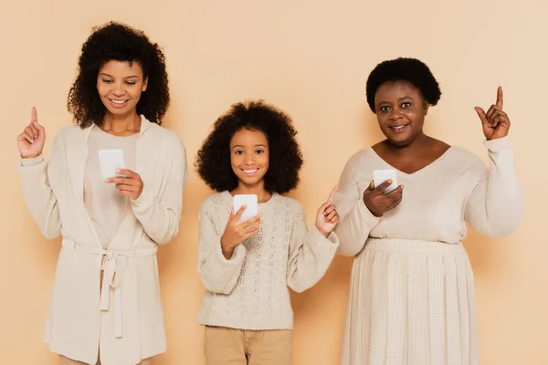 Afro-americana filha, neta e avó apontando com os dedos e segurando celulares em fundo bege — Fotografia de Stock
