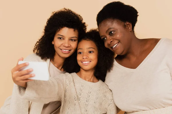 Smiling african american daughter, granddaughter and grandmother taking selfie on cellphone together on beige background — Stock Photo