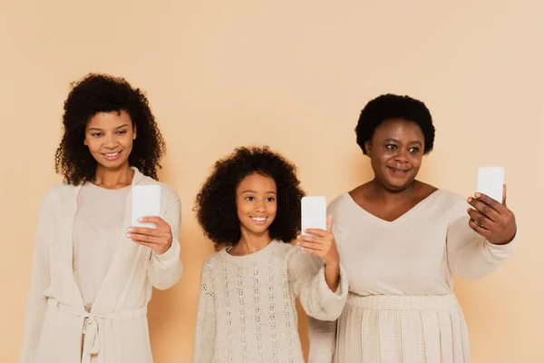 African american daughter, granddaughter and grandmother taking selfies on cellphones on beige background — Stock Photo