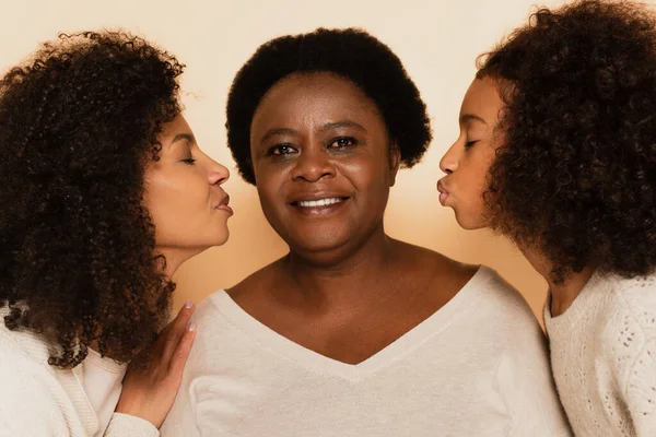 Afrikanisch-amerikanische Tochter und Enkelin küssen Großmutter mit geschlossenen Augen auf beigem Hintergrund — Stockfoto