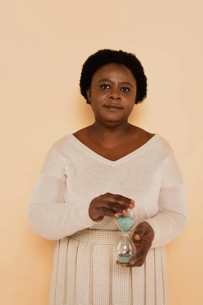 Mujer afroamericana seria sosteniendo vidrio de arena en las manos sobre fondo beige, pancarta — Stock Photo