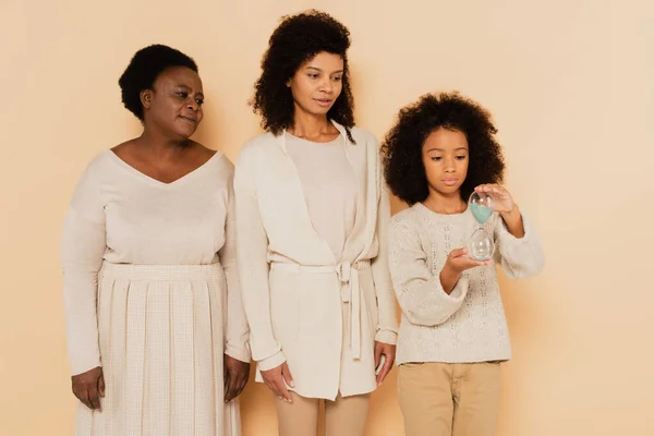 African american granddaughter holding sandglass near mother and grandmother on beige background — Stock Photo