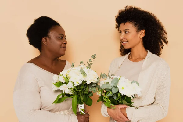 African american middle aged mother and adult daughter looking at each other with bouquets of flowers on beige background — Stock Photo