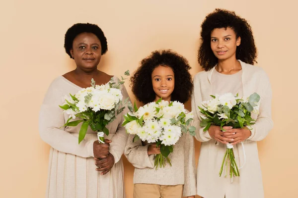 Happy family of african american grandmother, daughter and granddaughter with bouquets of flowers isolated on beige — Stock Photo