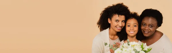 Família afro-americana feliz de mãe, filha e avó abraçando e segurando buquê de flores isoladas em bege, bandeira — Fotografia de Stock