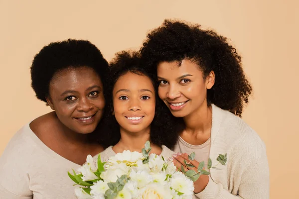 Glücklich afrikanisch-amerikanische Familie von Mutter, Tochter und Großmutter umarmen und halten Blumenstrauß isoliert auf beige — Stockfoto