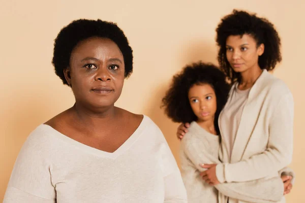 Afro-américaine mère embrassant avec sa fille et regardant grand-mère debout sérieusement sur fond beige — Photo de stock