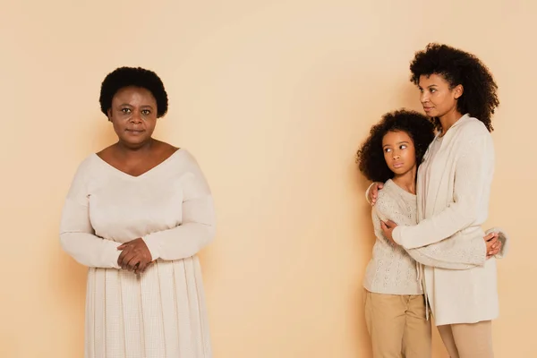 Afroamericano madre abbracciando con figlia e guardando la nonna seria su sfondo beige — Foto stock