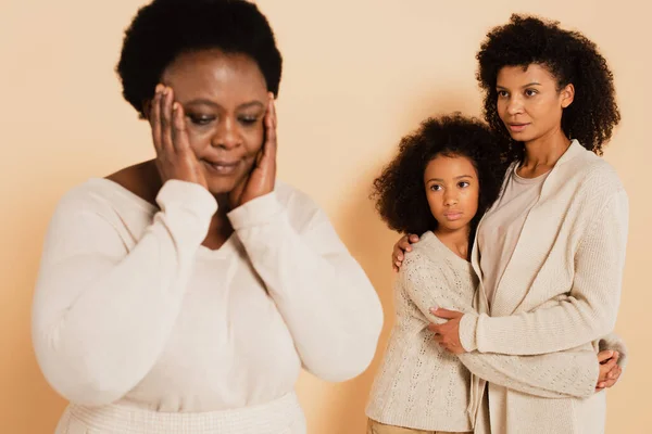 African american mother embracing with daughter and looking at disappointed grandmother with hands near head on beige background — Stock Photo