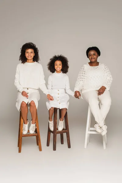Lächelnde afrikanisch-amerikanische Mutter, Tochter und Großmutter halten sich an den Händen und sitzen auf Hockern vor grauem Hintergrund — Stockfoto