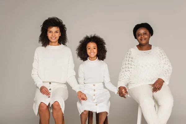 Happy african american mother, daughter and grandmother holding hands and sitting on stools on grey background — Stock Photo