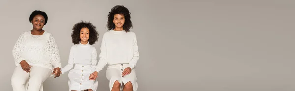 Heureuse mère afro-américaine, fille et grand-mère tenant la main et assise sur des tabourets sur fond gris, bannière — Photo de stock