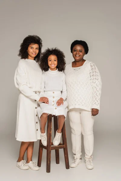 Happy african american mother and grandmother holding hands with granddaughter sitting on stool on grey background — Stock Photo