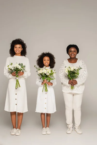 Vue pleine longueur de mère et grand-mère afro-américaine regardant sa petite-fille et tenant des bouquets de fleurs sur fond gris — Photo de stock