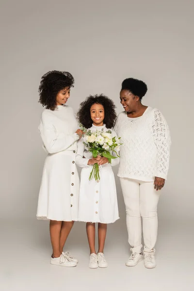 Souriant afro-américaine mère et grand-mère étreignant et regardant petite-fille avec bouquet de fleurs sur fond gris — Photo de stock