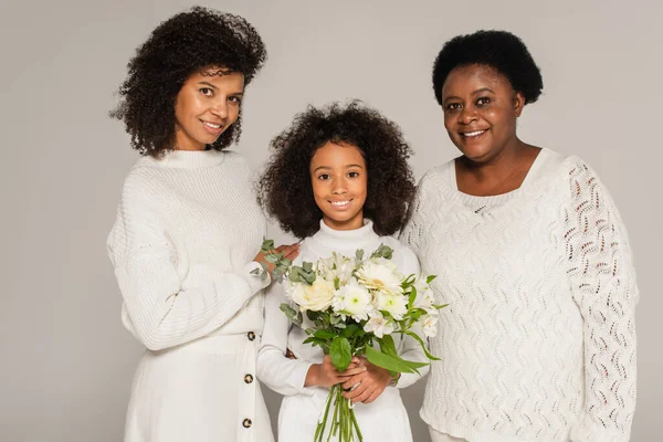 Souriant afro-américaine mère et grand-mère étreignant petite-fille souriante avec bouquet de fleurs isolées sur gris — Photo de stock