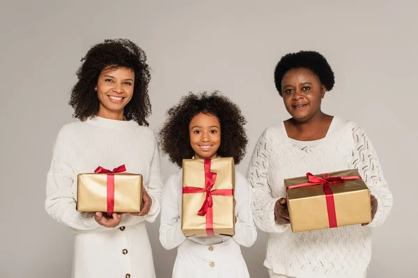 Улыбающаяся африканская американская мать, дочь и бабушка держат подарочные коробки изолированные на сером — стоковое фото