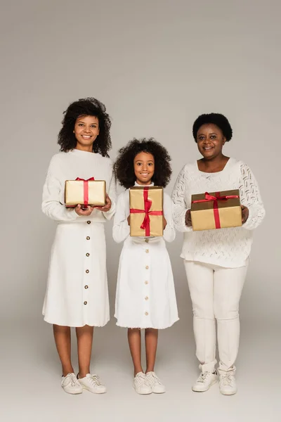 Полный снимок улыбающейся африканской матери, дочери и бабушки с подарочными коробками на сером фоне — стоковое фото