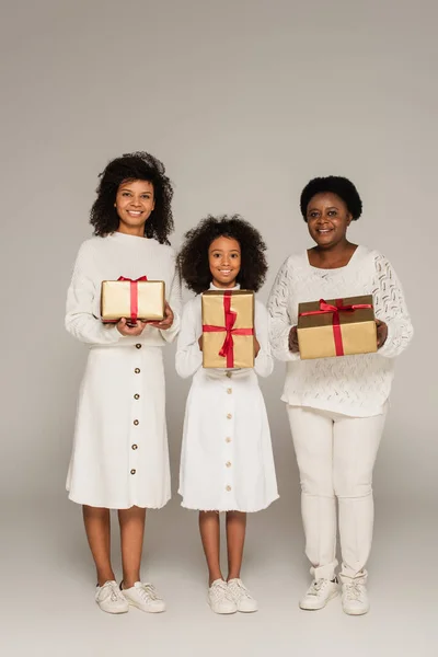 Full length shot of smiling african american mother, daughter and grandmother holding gift boxes on grey background — Stock Photo