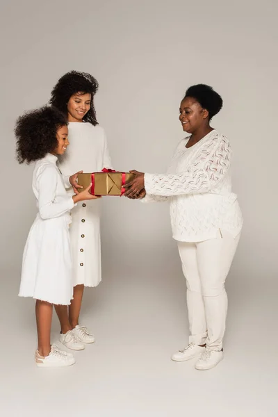 Ganzkörperaufnahme einer lächelnden afrikanisch-amerikanischen Großmutter, die Tochter und Enkelin eine Geschenkbox auf grauem Hintergrund präsentiert — Stockfoto